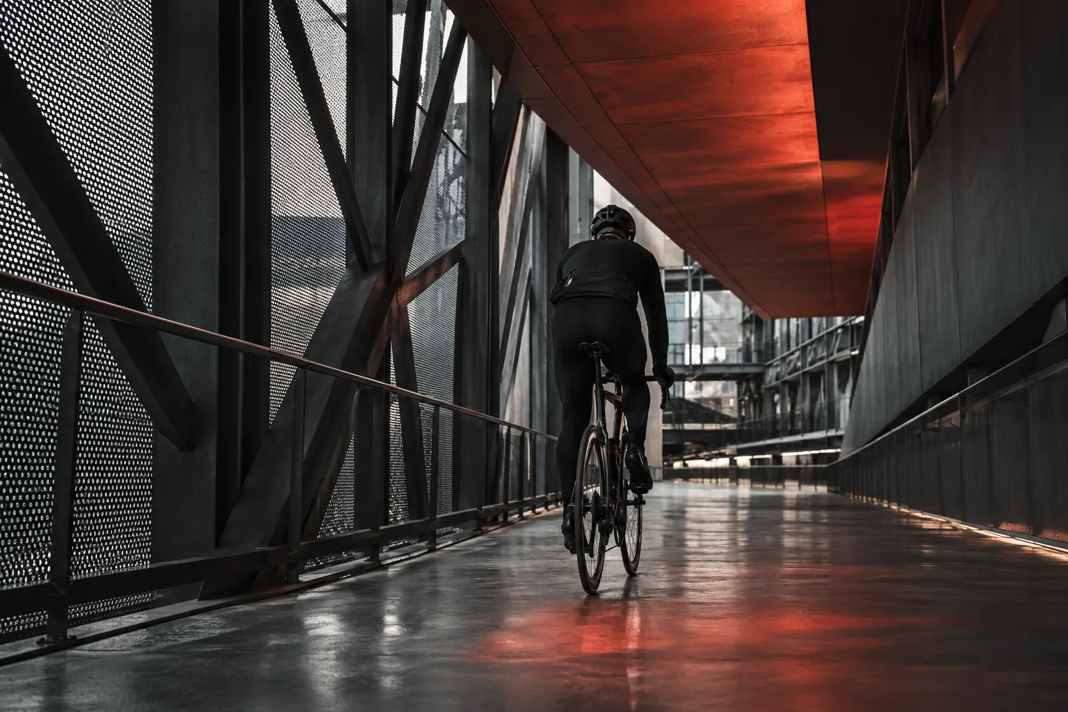 An adult cyclist in black technical clothing moving through a steel passage beneath a rust-red architectural plane.